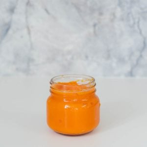 A glass jar of orange puree sitting in front of a grey marble splashback.