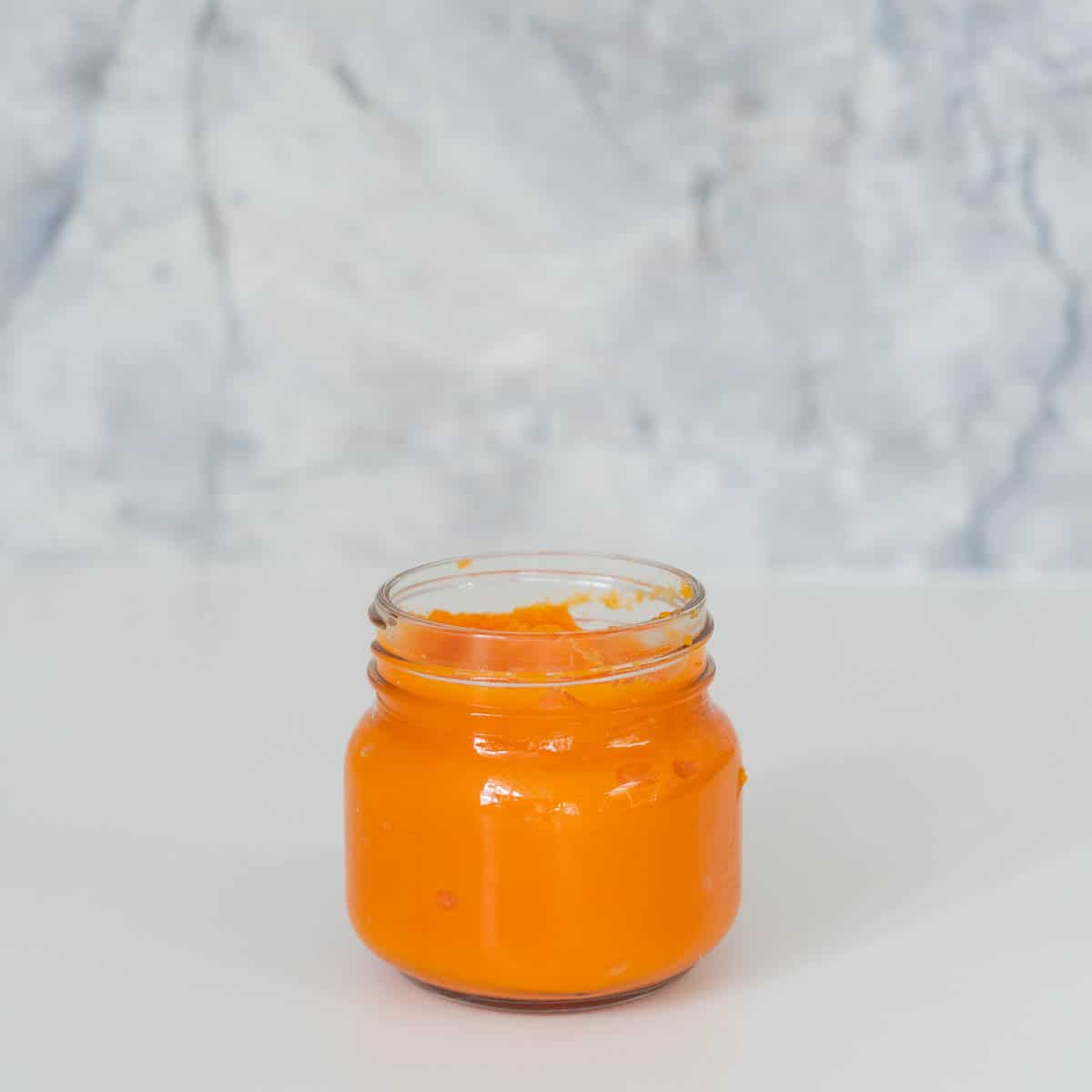 A glass jar of orange puree sitting in front of a grey marble splashback.