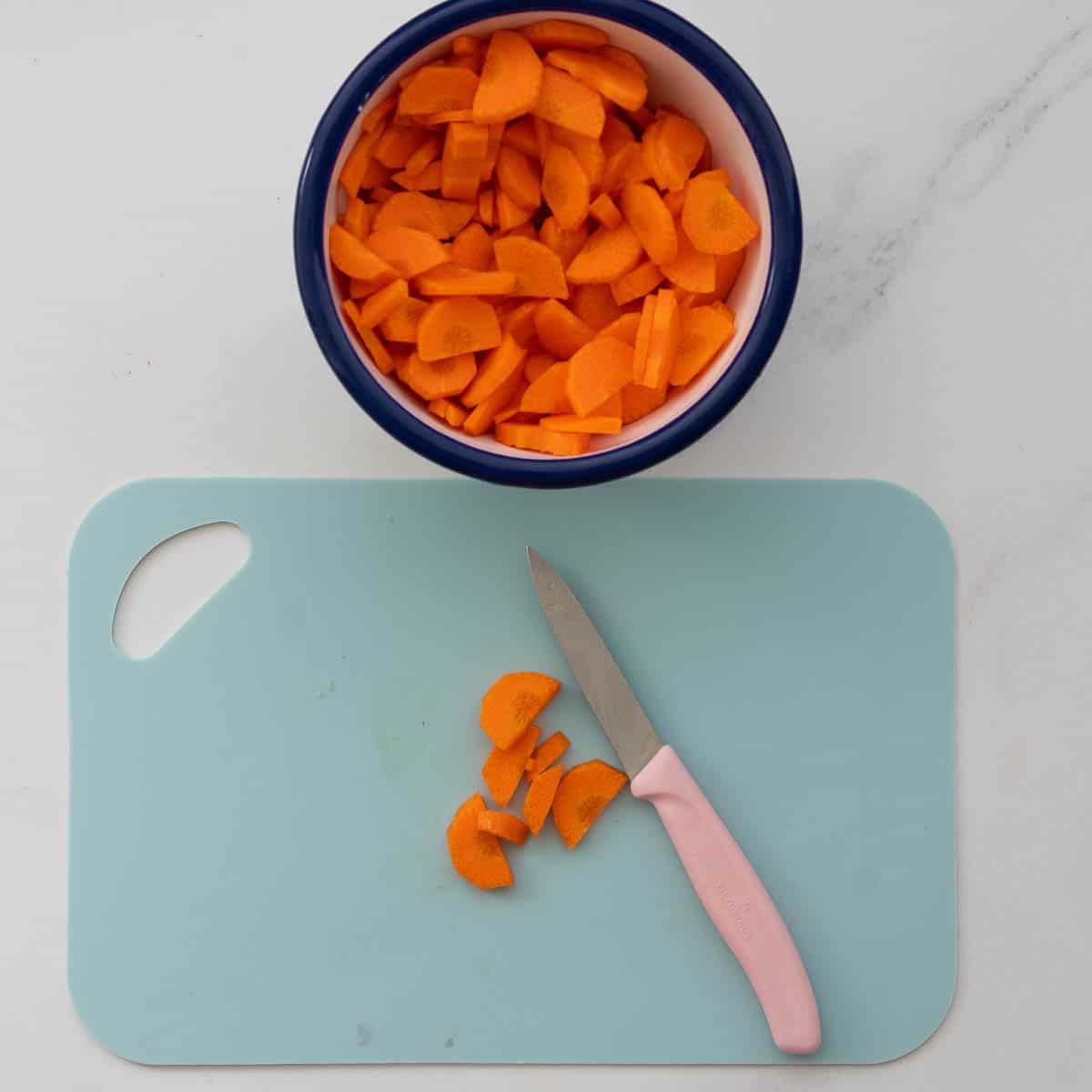 A white enamel bowl filled with slices of carrot next to a light blue chopping board and pink handled paring knife.