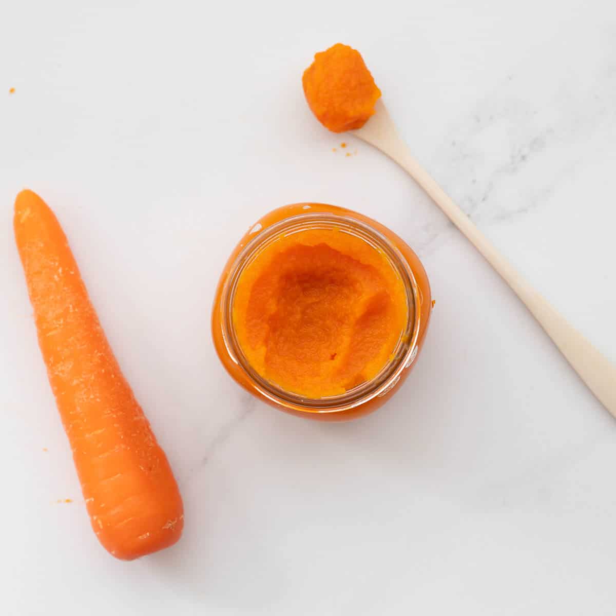 A jar of orange carrot puree on a bench next to a carrot  and a bamboo spoon loaded with the puree.
