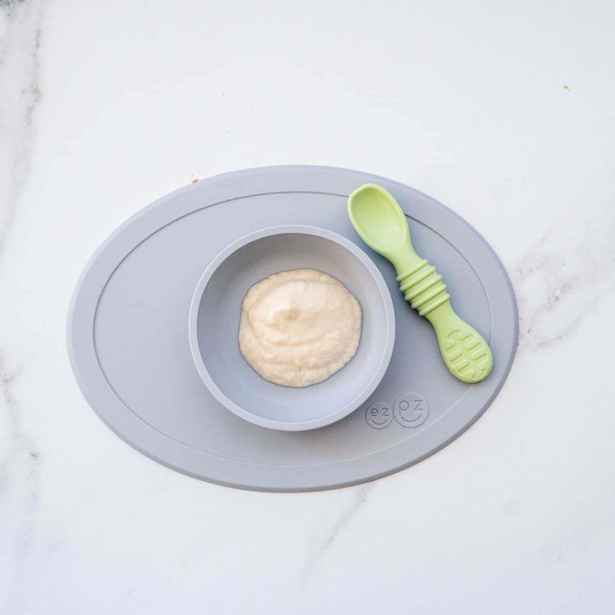 A grey silicone baby plate filled with white puree, with a pale green baby spoon lying to the side.