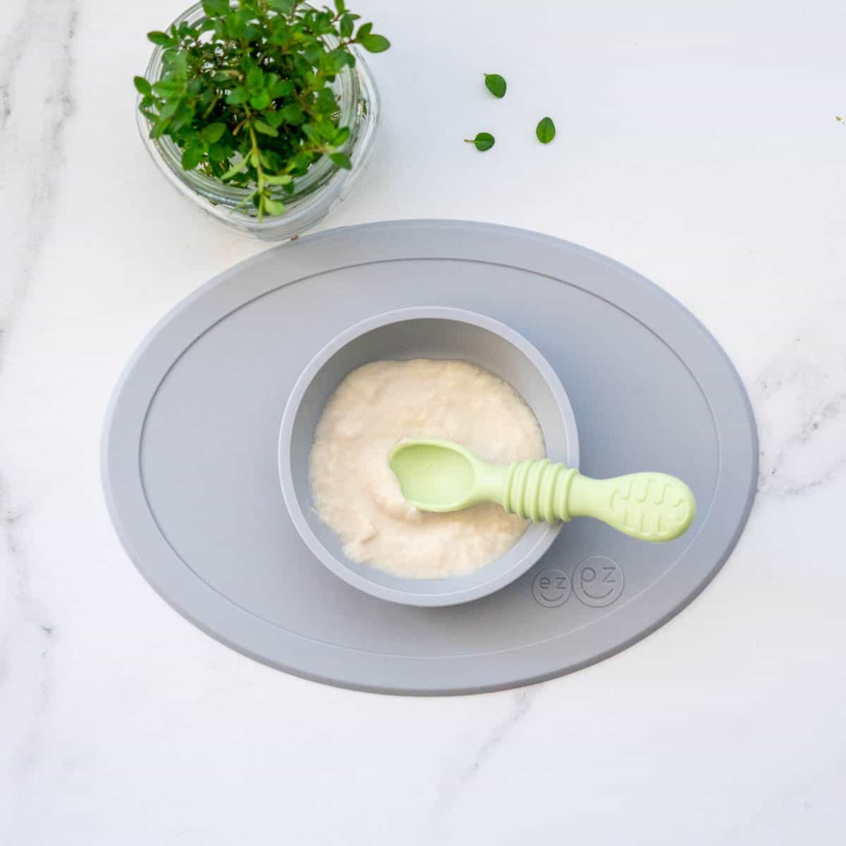 A grey silicone baby plate with pale green spoon filled with chicken puree next to a thyme plant.
