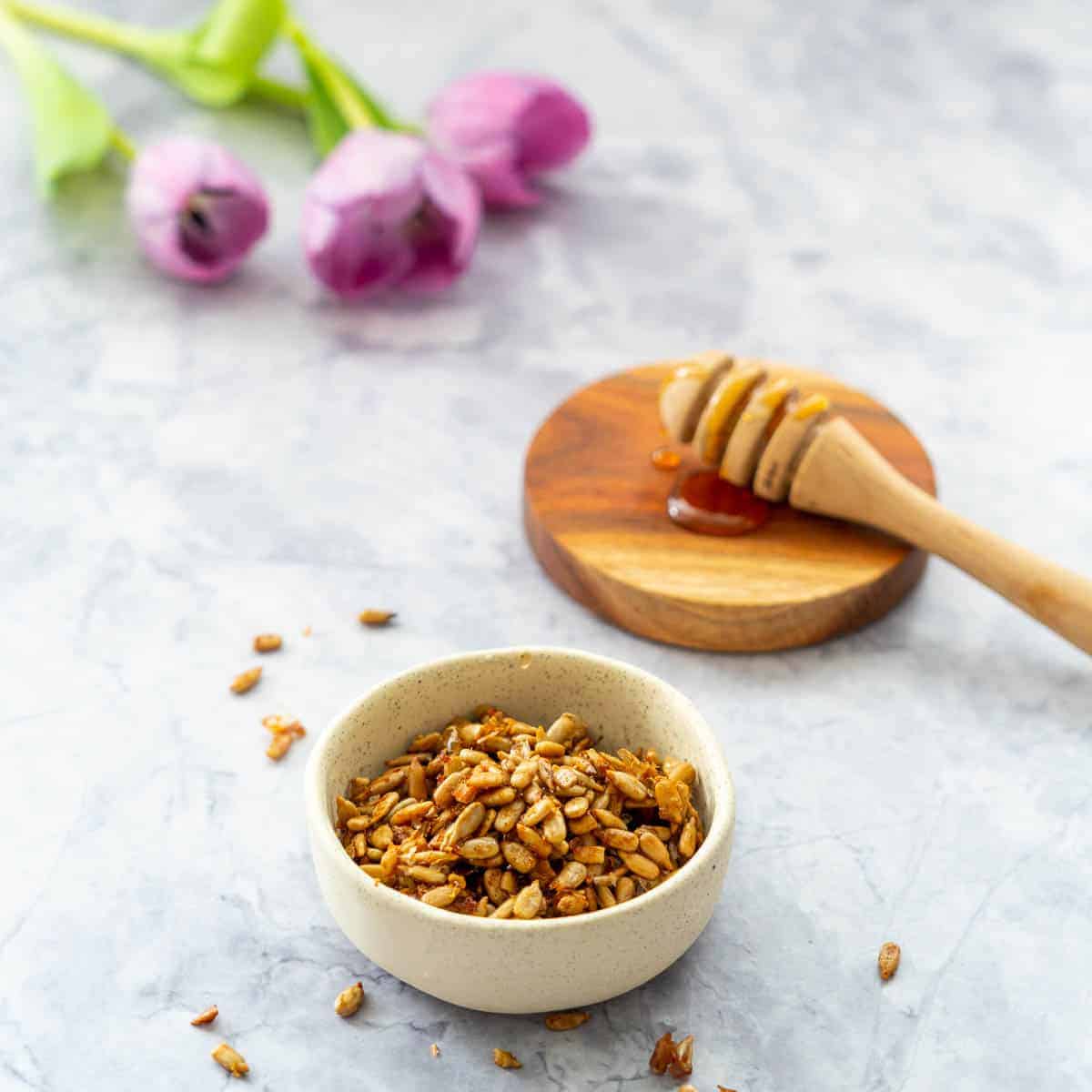 Faded in the background are some purple tulips beside the honey dipper stick on a wooden board. In front an oatmeal colour bowl with roasted seeds