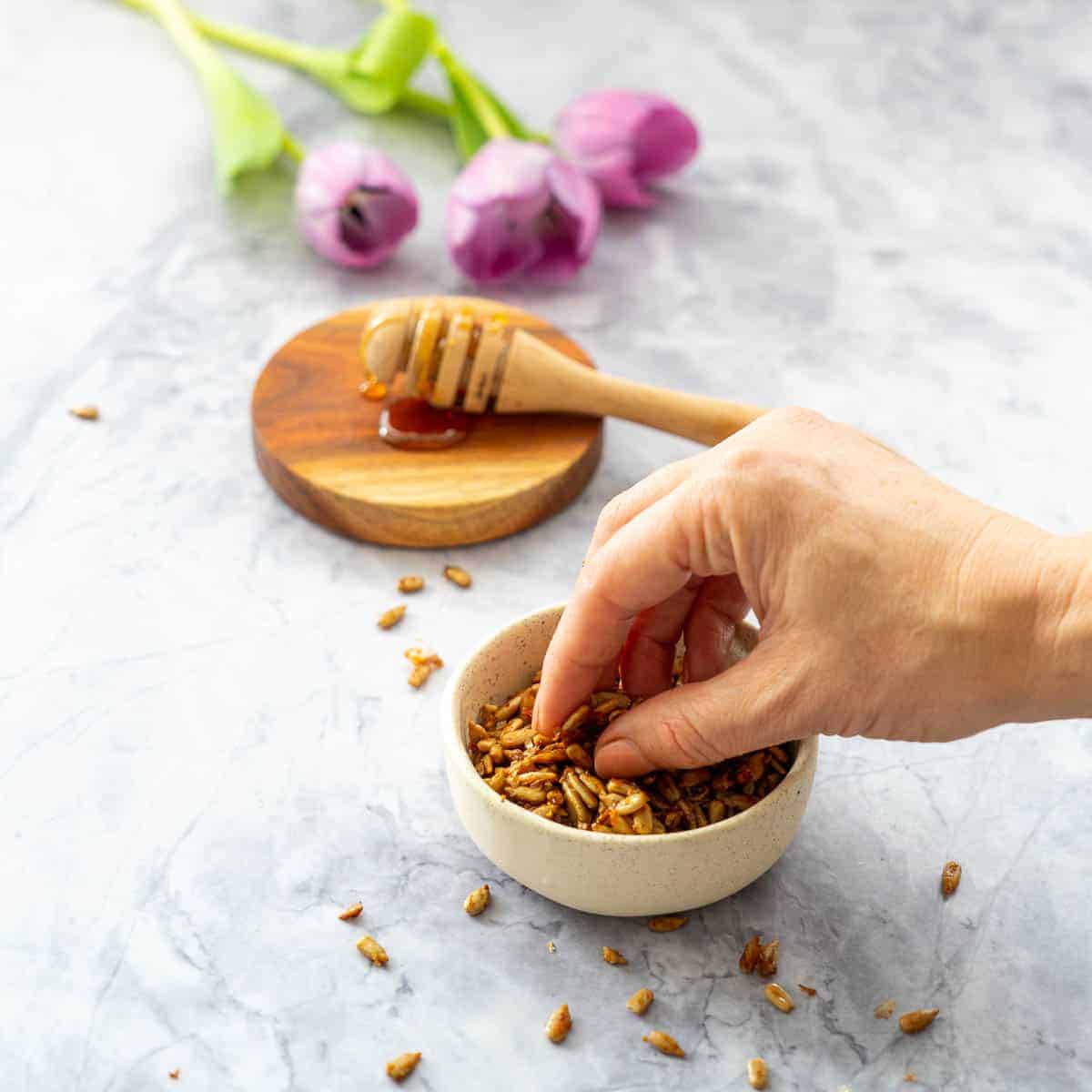 Faded in the background are some purple tulips beside the honey dipper stick on a wooden board. In front is a small oatmeal coloured bowl with roasted seeds with a hand pinching some.