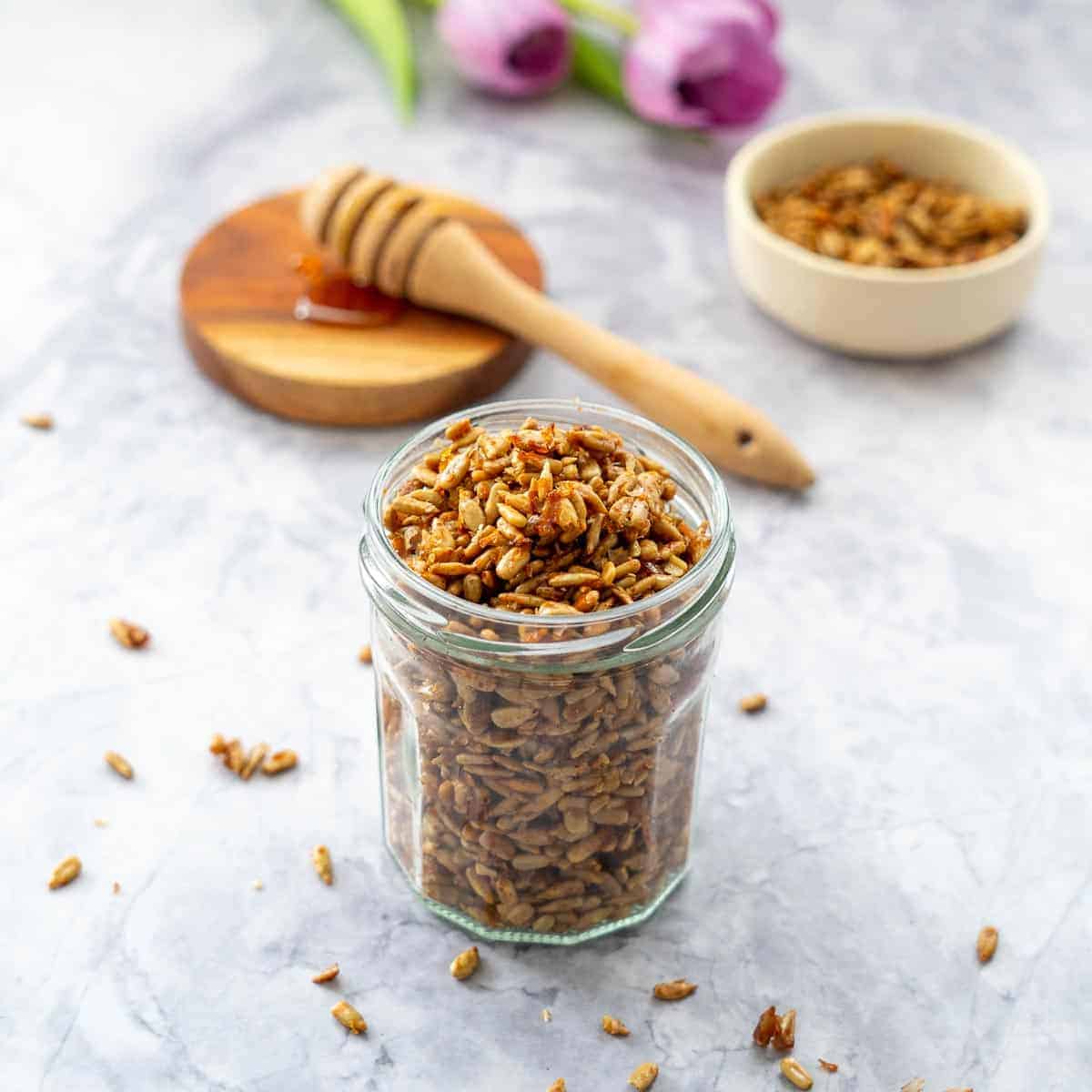 A clear jar filled with honey roasted sunflower seeds. In the background is a honey dipper stick dripping onto a wooden coaster with a side of roasted seeds