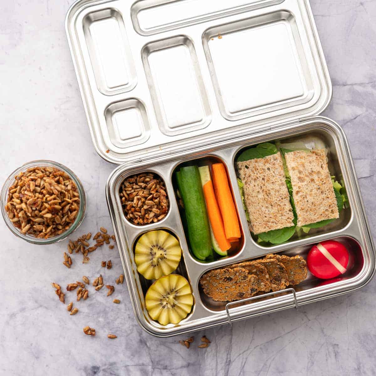 A stainless steal lunch box filled with the roasted seeds along with a kiwifruit, cucumber and carrot slides, cheese round and crackers and a brown bread sandwich.