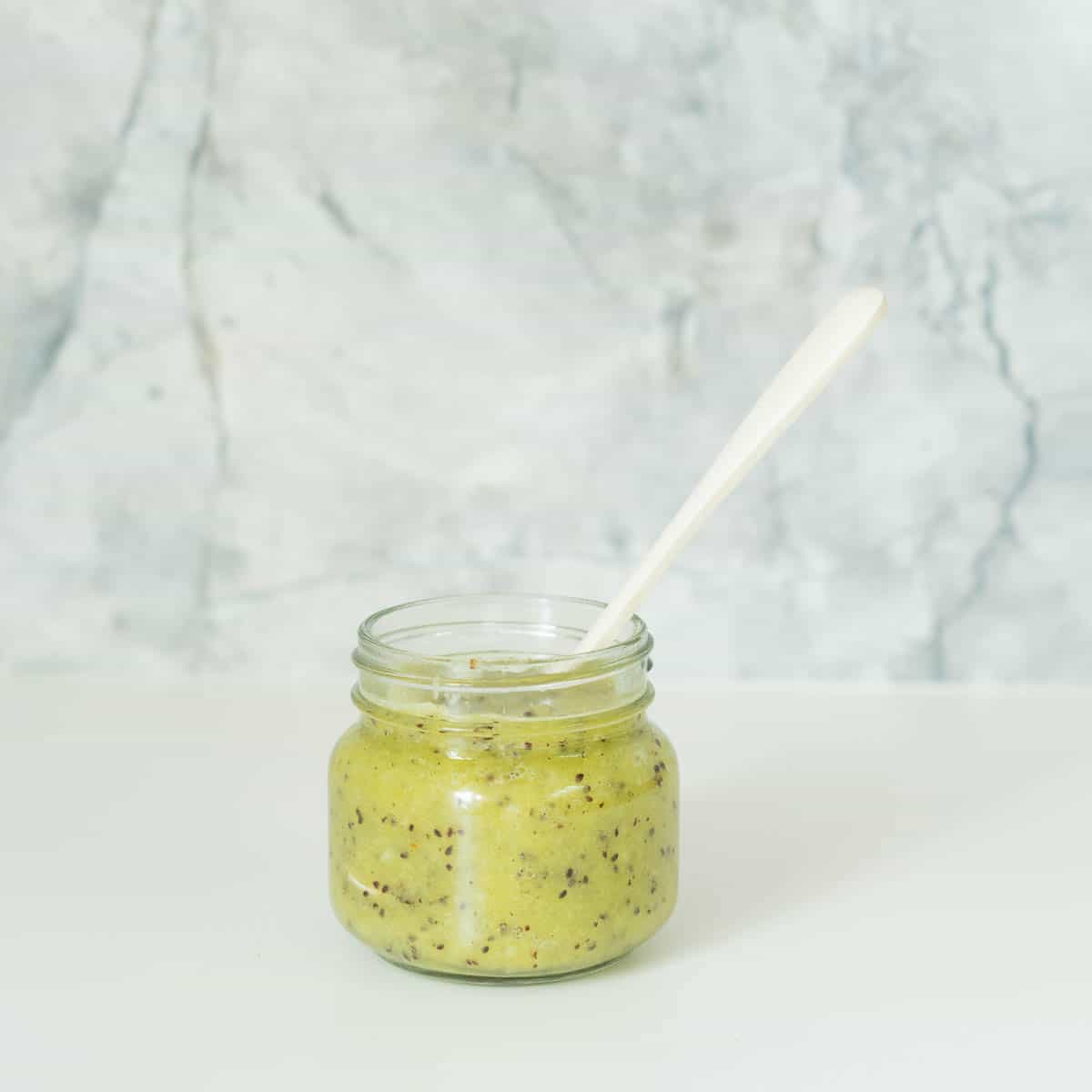 A small glass jar filled with kiwifruit puree in front of a grey marble splashback.