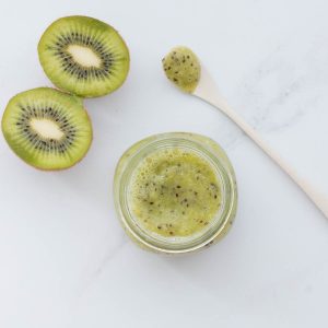 A jar of green kiwi puree on a bench next to slices of kiwifruit and a bamboo spoon.