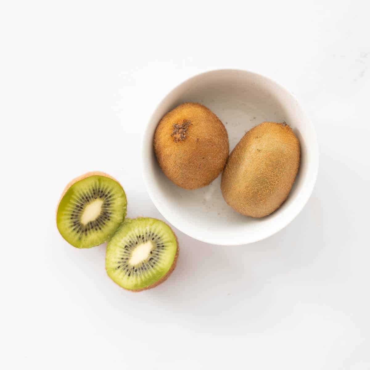 Two kiwifruit in a small ceramic bowl, next to a kiwifruit that has been sliced in half.