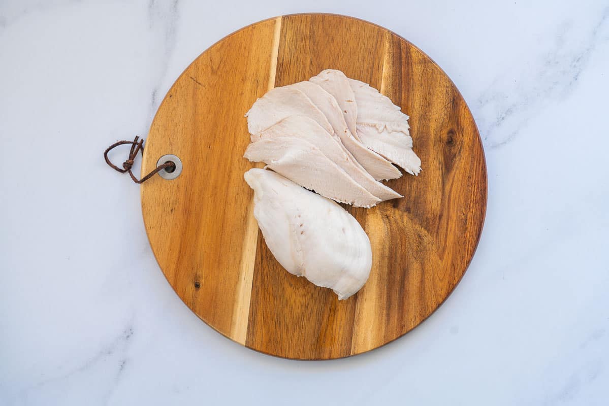 One whole poached chiken breast and one sliced chicken breast on a round wooden chopping board. 