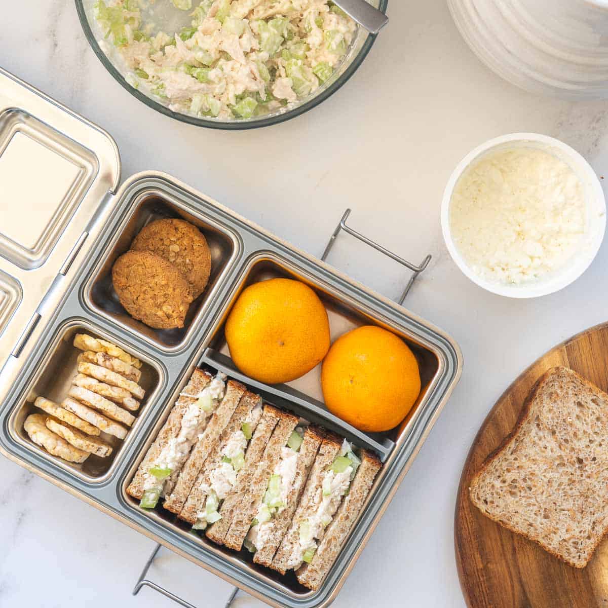 A stainless steel lunchbox packed with fruit, crackers and shredded chicken sandwiches. 
