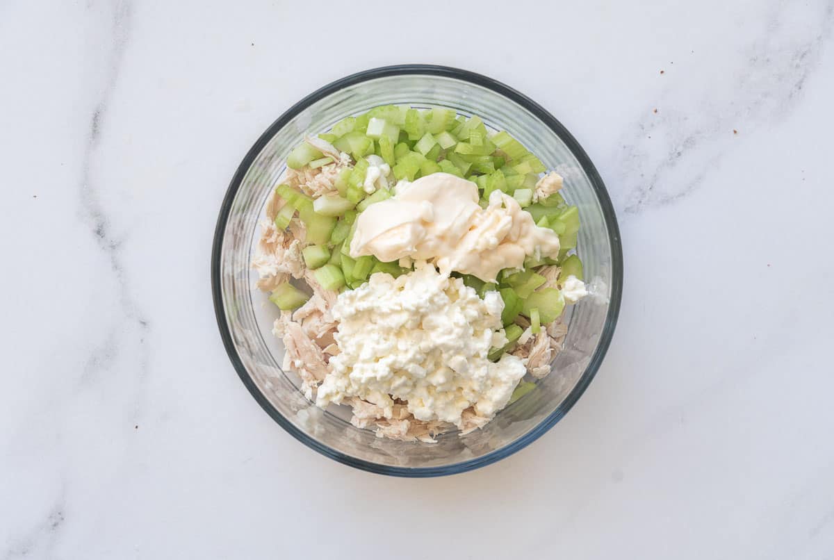 Shredded chicken, cottage cheese, diced celery and mayonnaise in a glass mixing bowl.