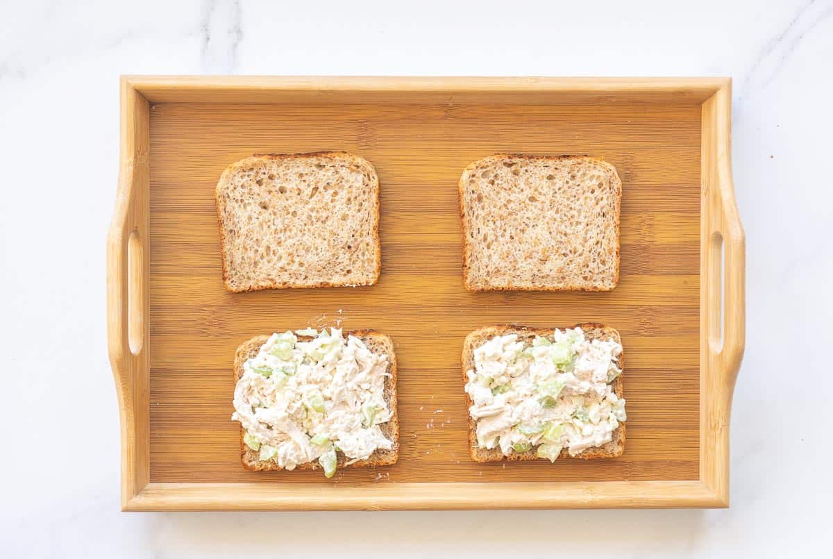 Four slices of bread on a wooden tray, two of them spread with chicken salad.