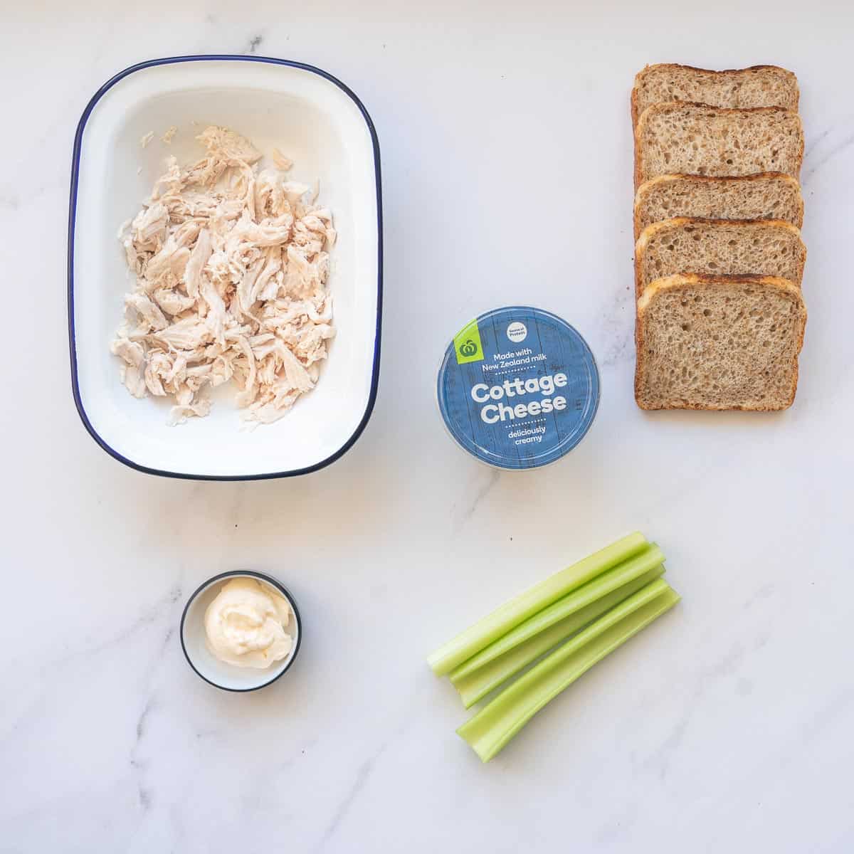 A dish of shredded chicken, small bowl of mayonnaise, celery sticks cottage cheese and slices of bread laid out on a bench top.