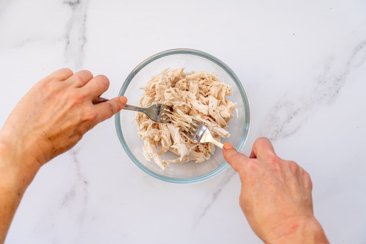 A glass bowl of poached chicken being shredded with the two fork method.