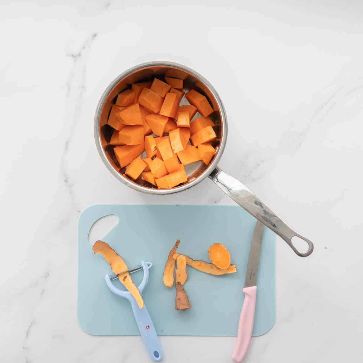 On the bench you see a pot with peeled and chopped sweet potato in front of a blue chopping board with a blue peeler and peel and a pink vege knife