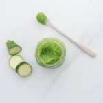 A jar of green vegetable puree on a bench next to slices of zucchini and a bamboo spoon.