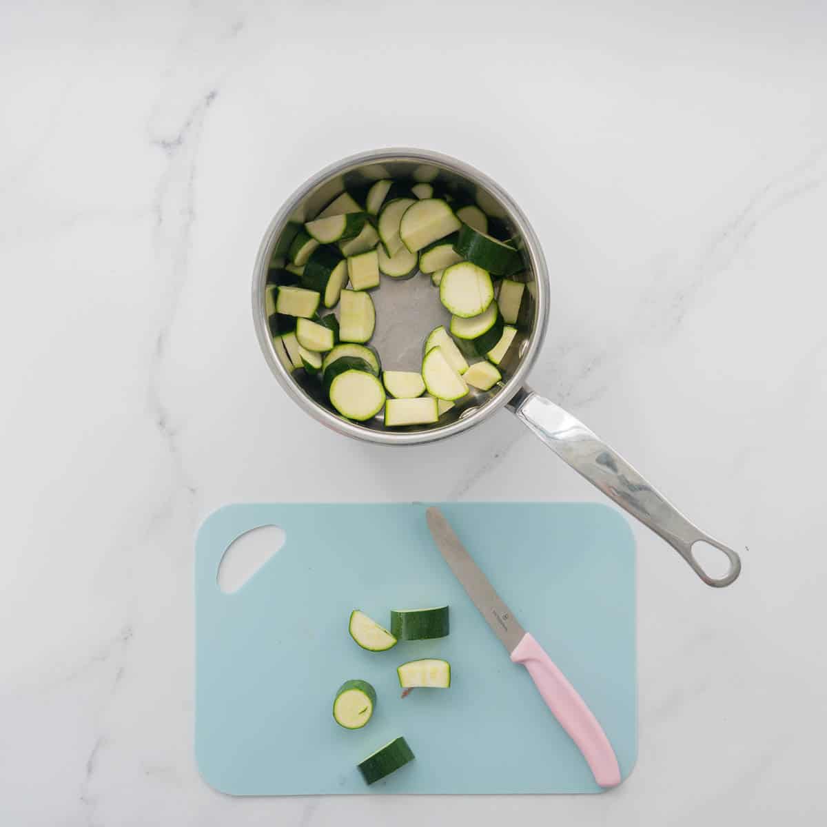 Slices of zucchini in a saucepan, next to a light blue chopping board and a pink handled vegetable knife. 