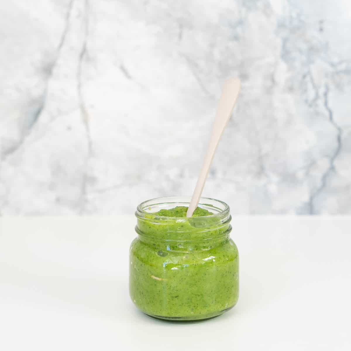 A glass jar of green zucchini puree sitting in front of a grey marble splashback.