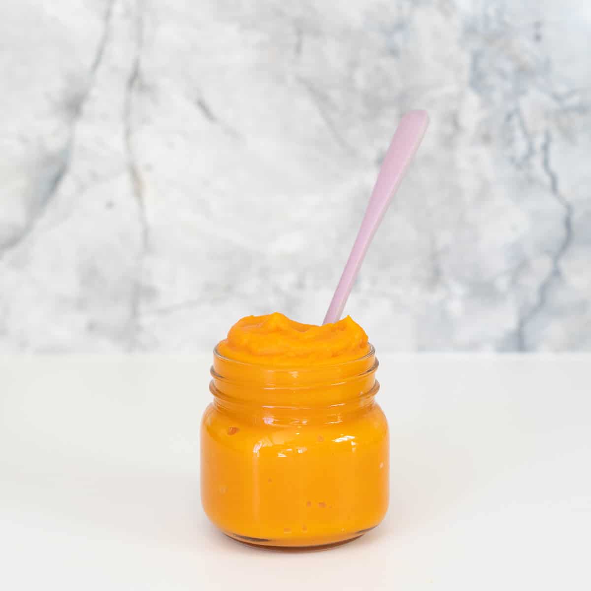 A small glass jar filled with orange baby puree and a purple wooden baby spoon.