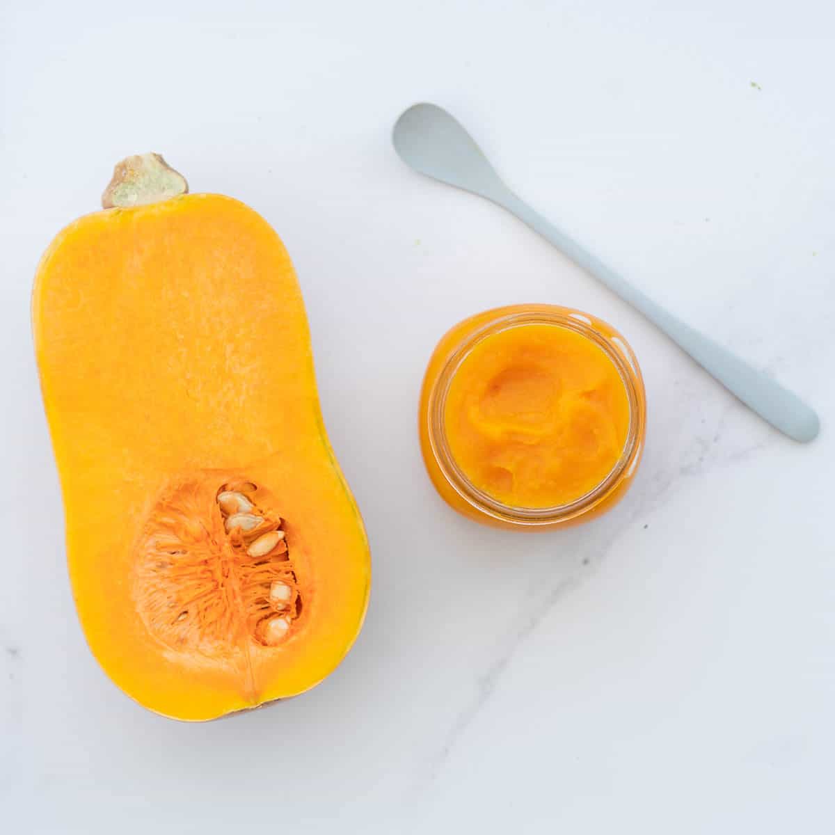 A jar of orange puree next to half a butternut squash and a blue baby spoon on a bench top.