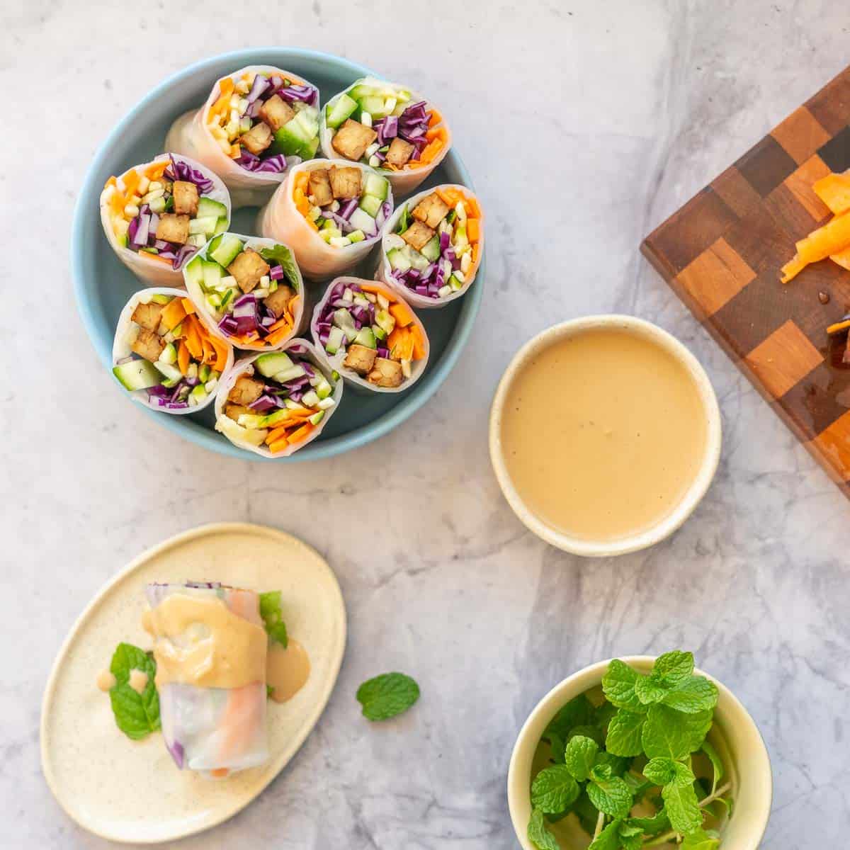 A platter of rice paper rolls, on a marble bench top with a dish of satay sauce, a side plate and fresh herbs.