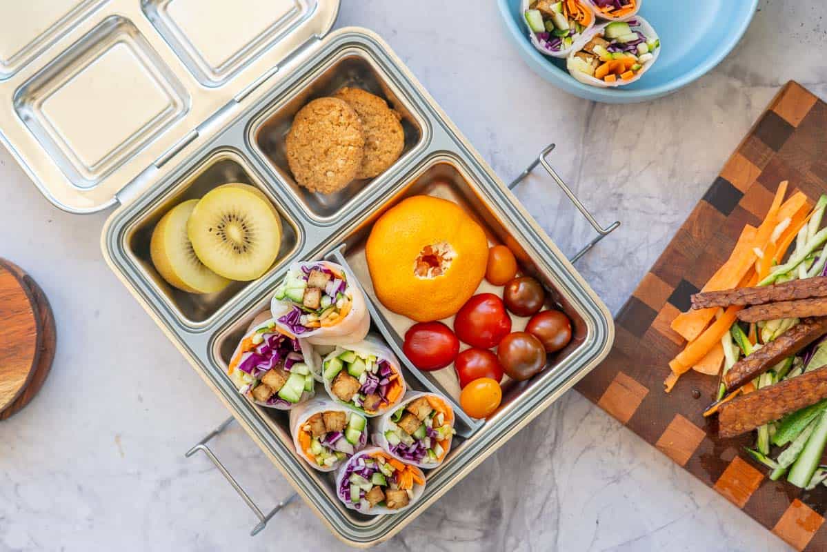 A stainless steel lunch box packed with rice paper rolls, fruit, vegetables and cookies.