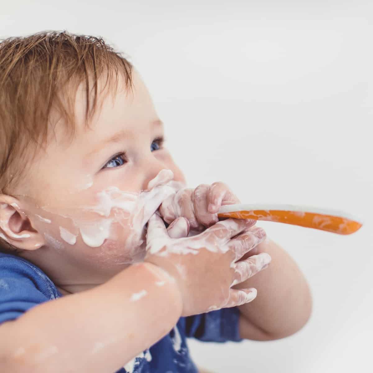 A baby messily spoon feeding themselves yogurt. 