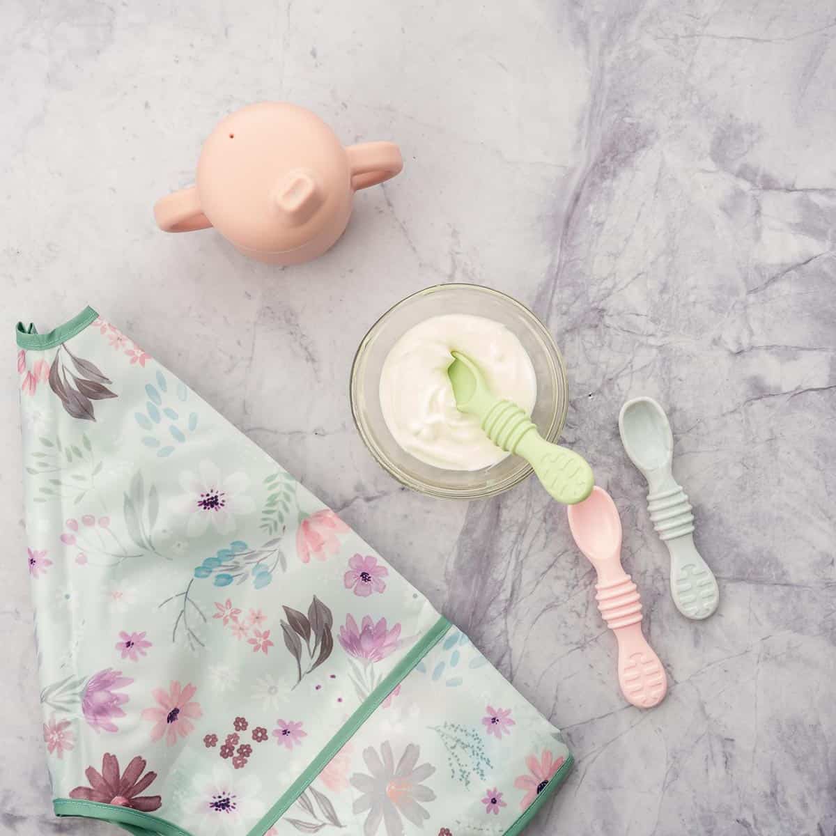 A small bowl of yogurt with baby spoons, a pink sippy cup and a floral bib.