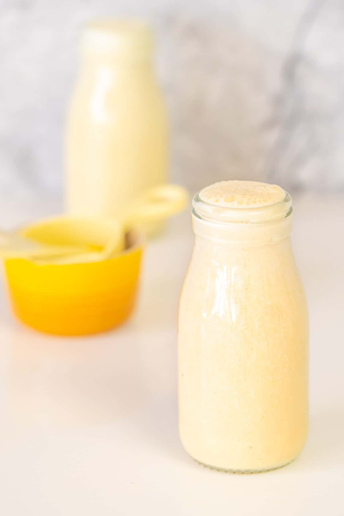 A small glass milk bottle filled with banana milk next to a yellow ramekin and yellow measuring cup. 