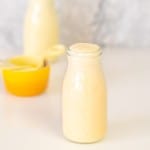 A small glass milk bottle filled with banana milk next to a yellow ramekin and yellow measuring cup.