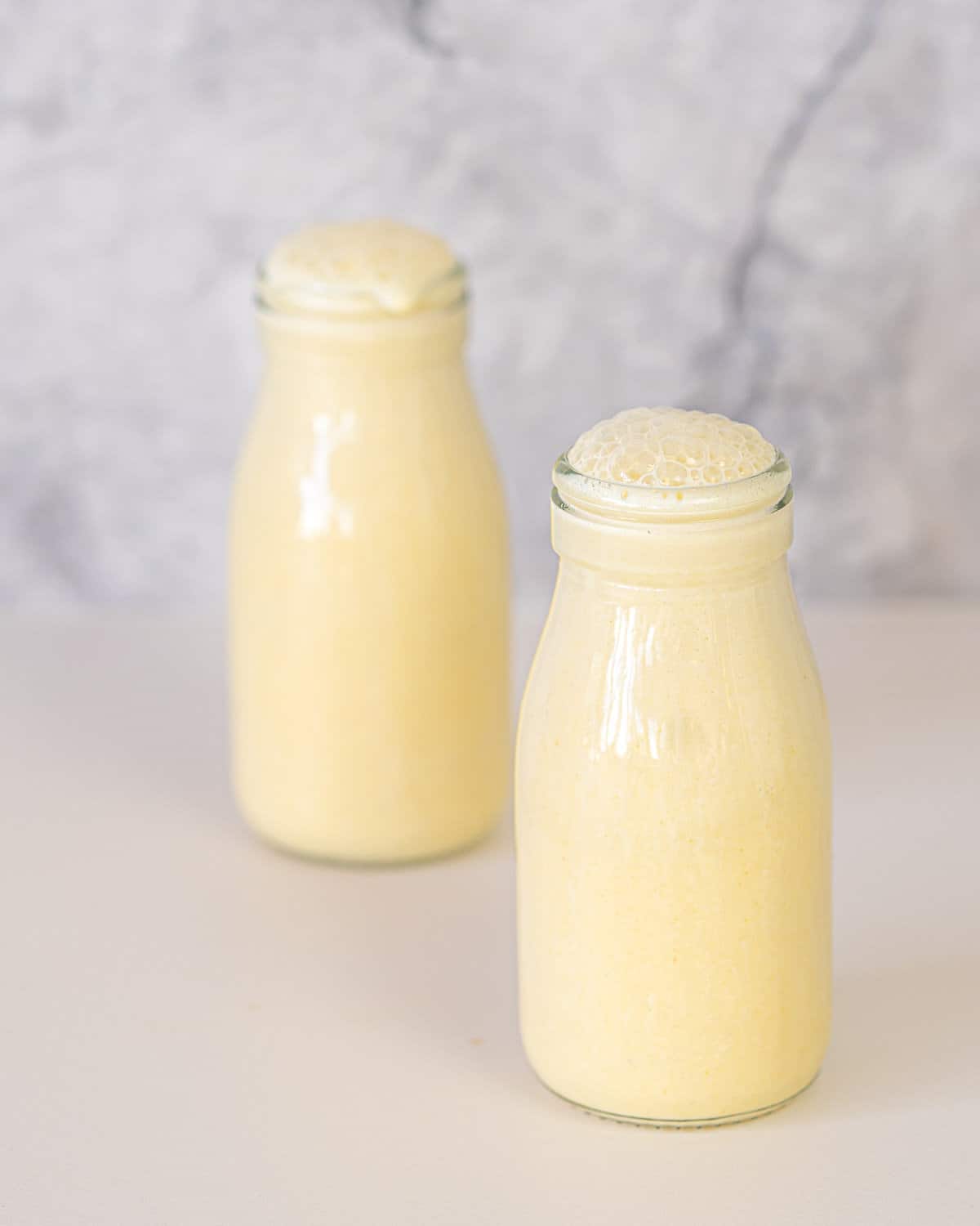 Two bottles of frothy yellow coloured milk sitting on a bench top in front of a marble spalshback. 