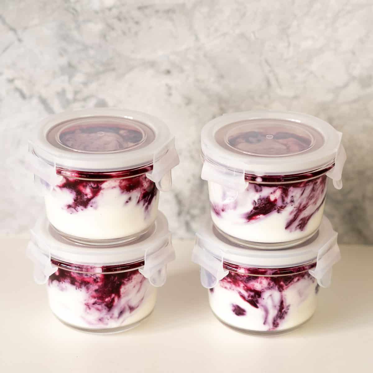 Four glass jars with white lids, filled with layers of blueberries and yogurt on a bench top.
