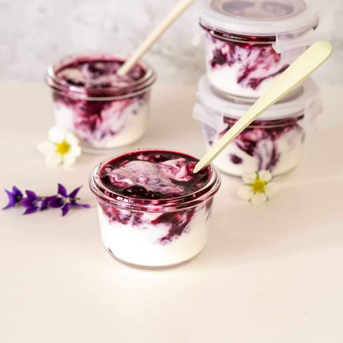 Blueberry yogurt in small glass jars on a bench top scattered with white and purple flowers.
