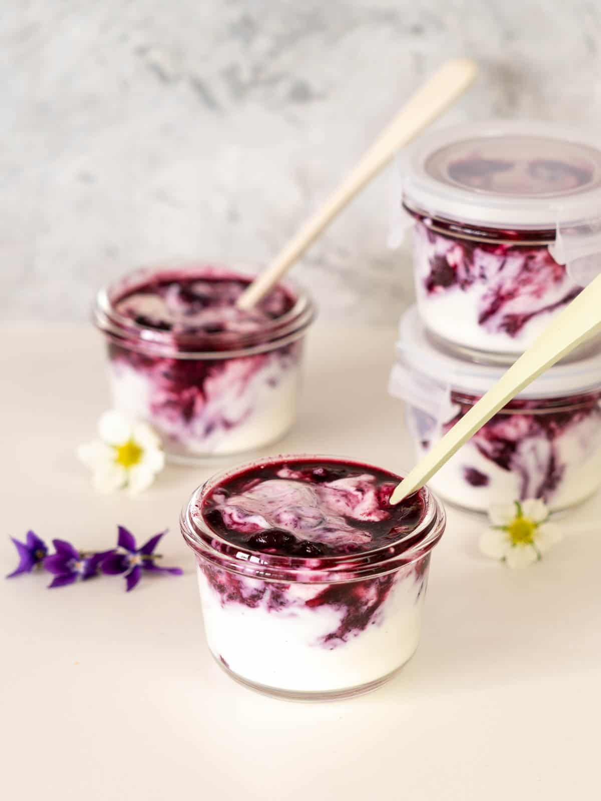 Blueberry yogurt in small glass jars on a bench top scattered with white and purple flowers.