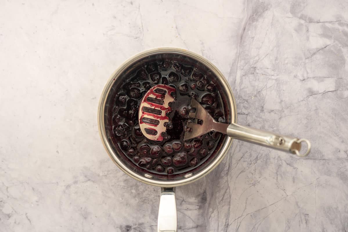 A saucepan of blueberry compote with a potato masher resting in it.