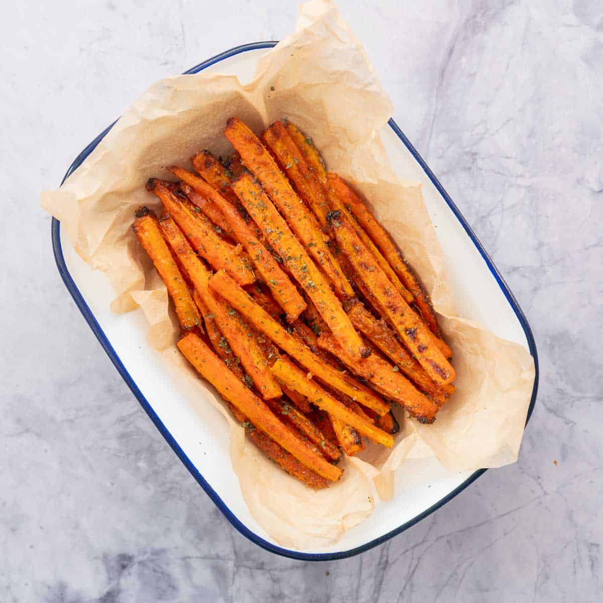 Carrot fries sprinkled with salt and parsley flakes in a white enamel serving bowl lined with crinkled parchment paper. 