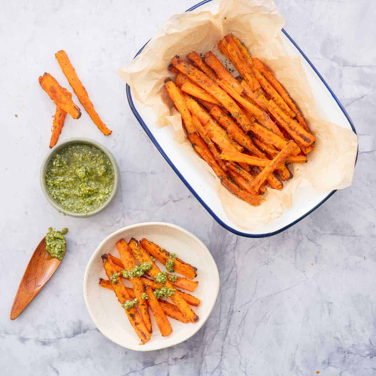 A serving dish filled with carrot fries next to a bowl of pesto sauce, and a side plate of carrot fries drizzled with the green pesto.