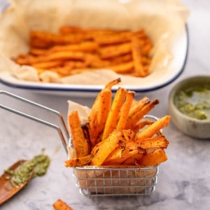 A small metal basket filled with carrot fries on a bench next to a spoonful of green pesto.