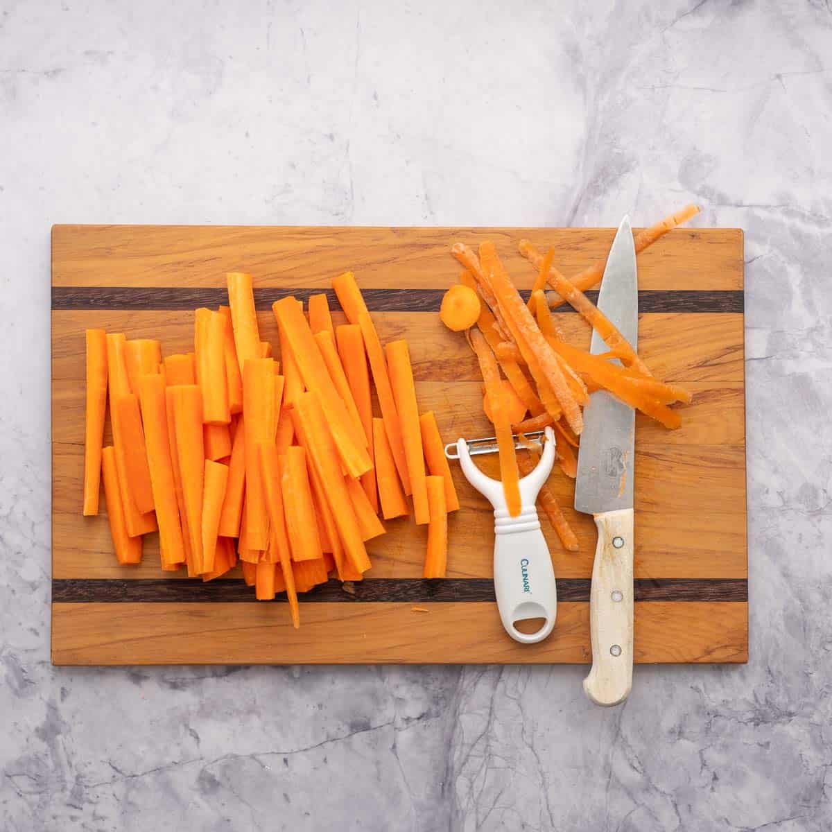 Carrot sticks on a wooden chopping board with a knife and vegetable peeler.