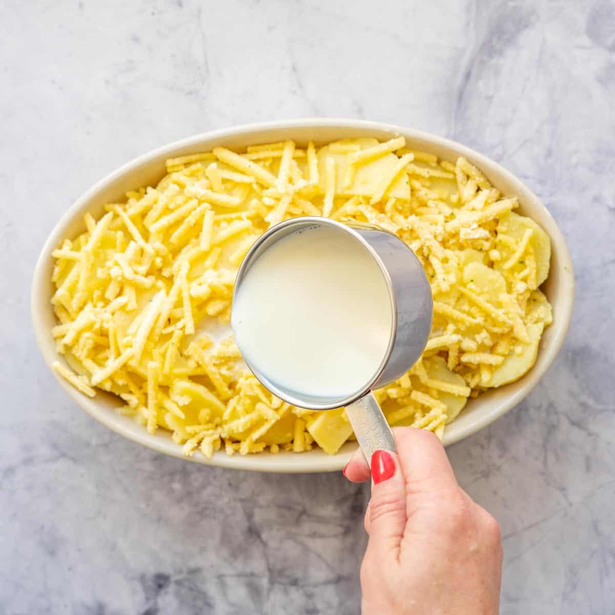 A measuring cup of milk being poured over a baking dish of potatoes and cheese.
