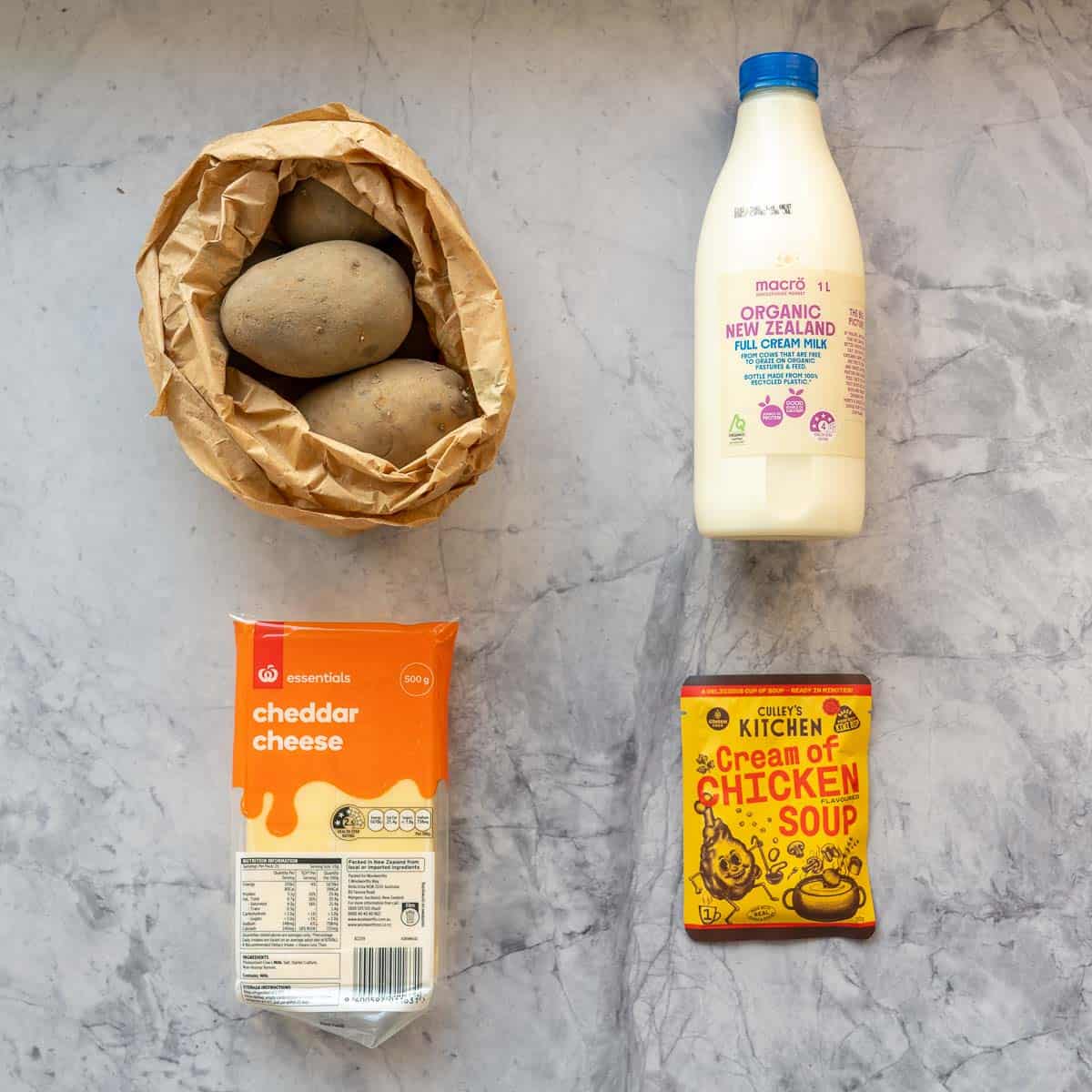 A brown paper bag of potatoes, bottle of milk, block of cheese and a packet of soup mix laid out on a bench top.