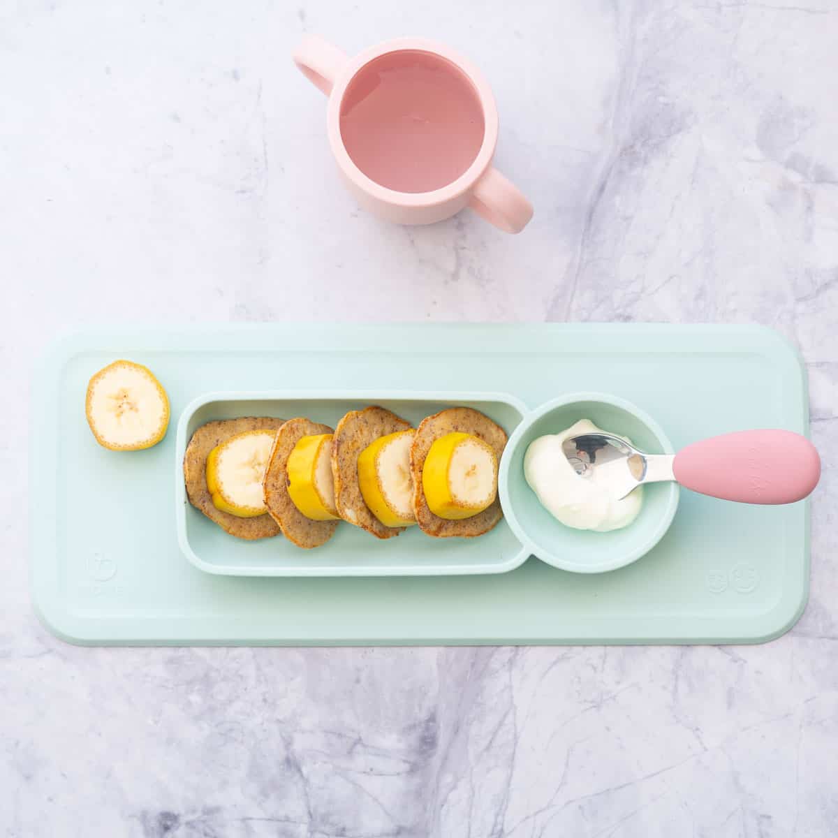 Banana slices, pancakes and yogurt on a mint green baby plate with a pink handled spoon and pink sippy cup. 