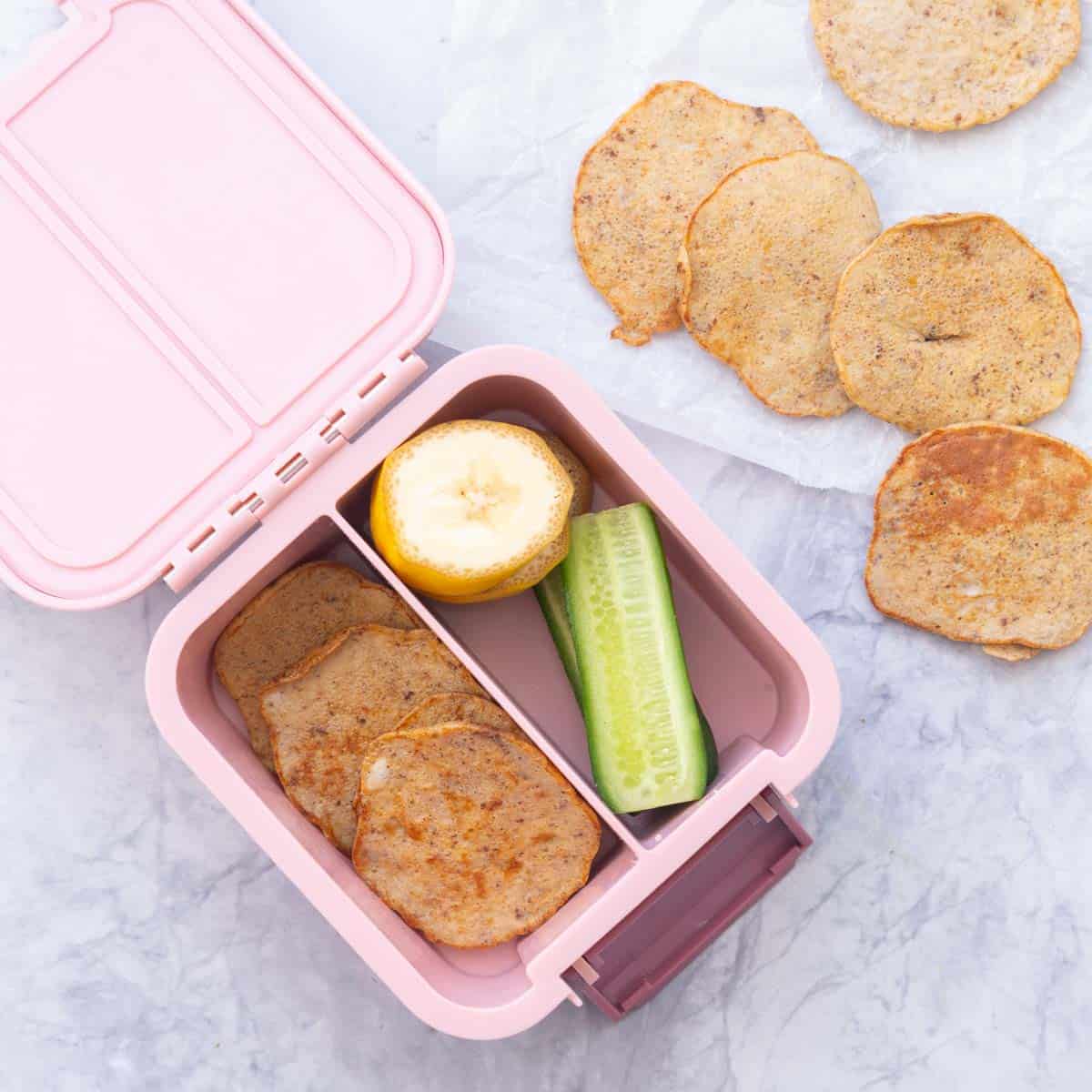 a pink bento box packed with pancakes, banana slices and cucumber sticks. 