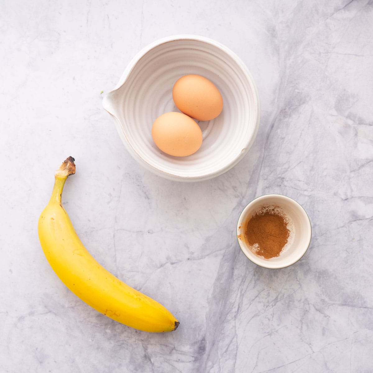 eggs, banana and a small dish of cinnamon on a marble bench top.
