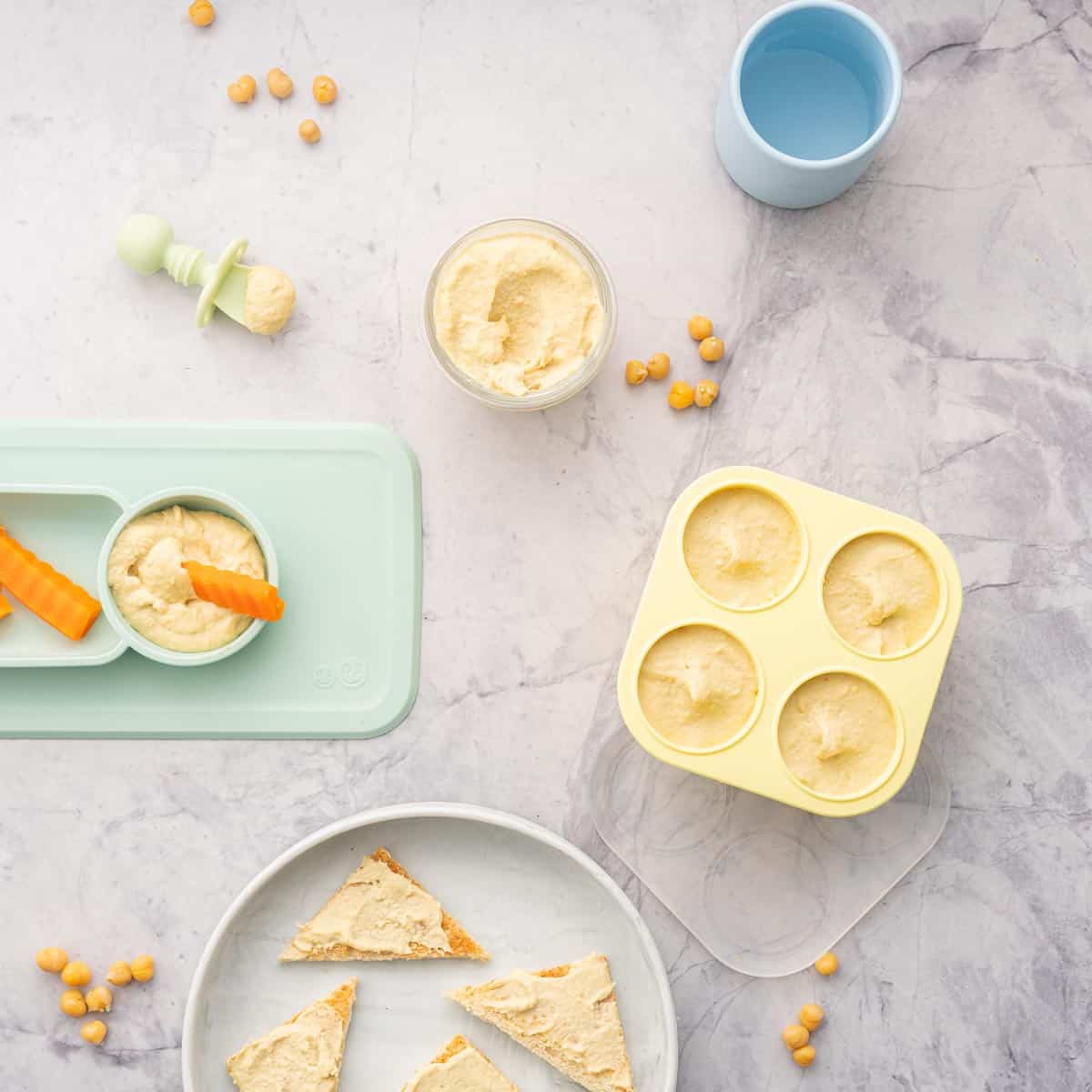 Yellow muffin cases filled with baby hummus sitting next to a ramekin of hummus and a baby plate with carrots dunked into hummus, chickpeas scattered on the bench next to a baby spoon and cup of water and a plate with toast with hummus spread 