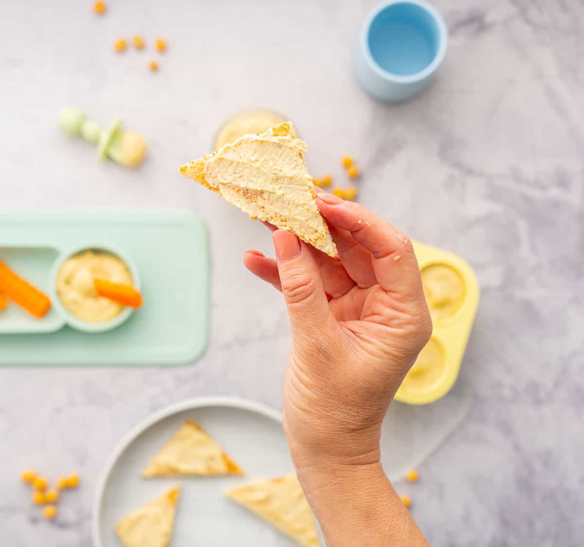 A hand holding up a slice of toast with hummus spread on it hovering above a yellow muffin case filled with baby hummus sitting next to a ramekin of hummus and a baby plate with carrots dunked into hummus, chickpeas scattered on the bench.