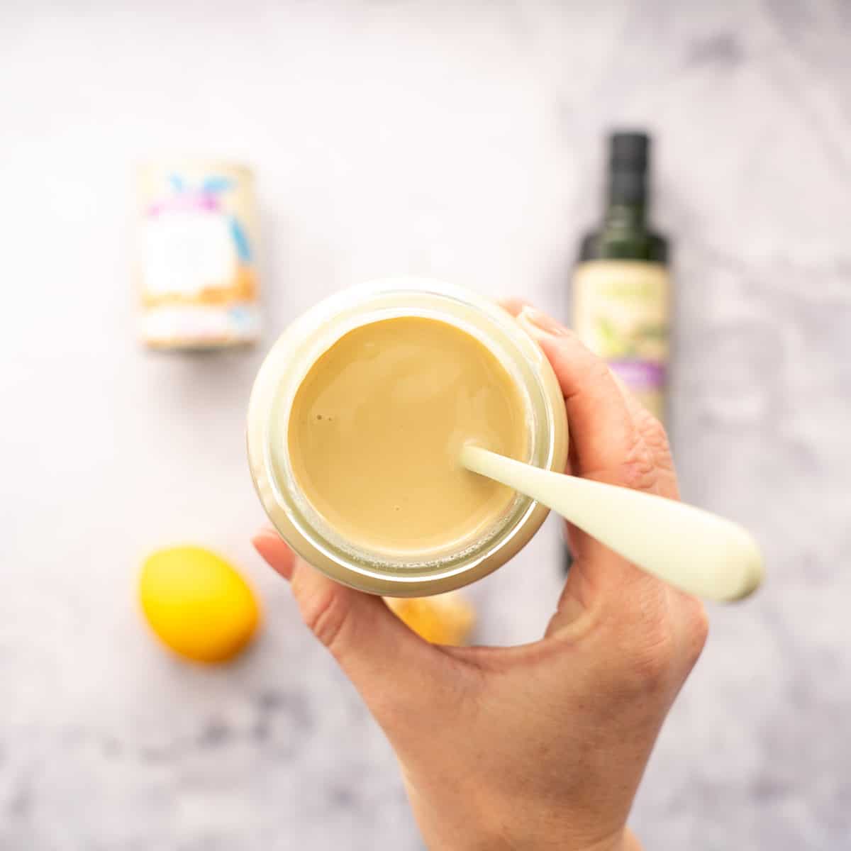 a hand holding up a jar of tahini with a spoon inside of it, hovering above the bench with the laid out ingredients below it. 
