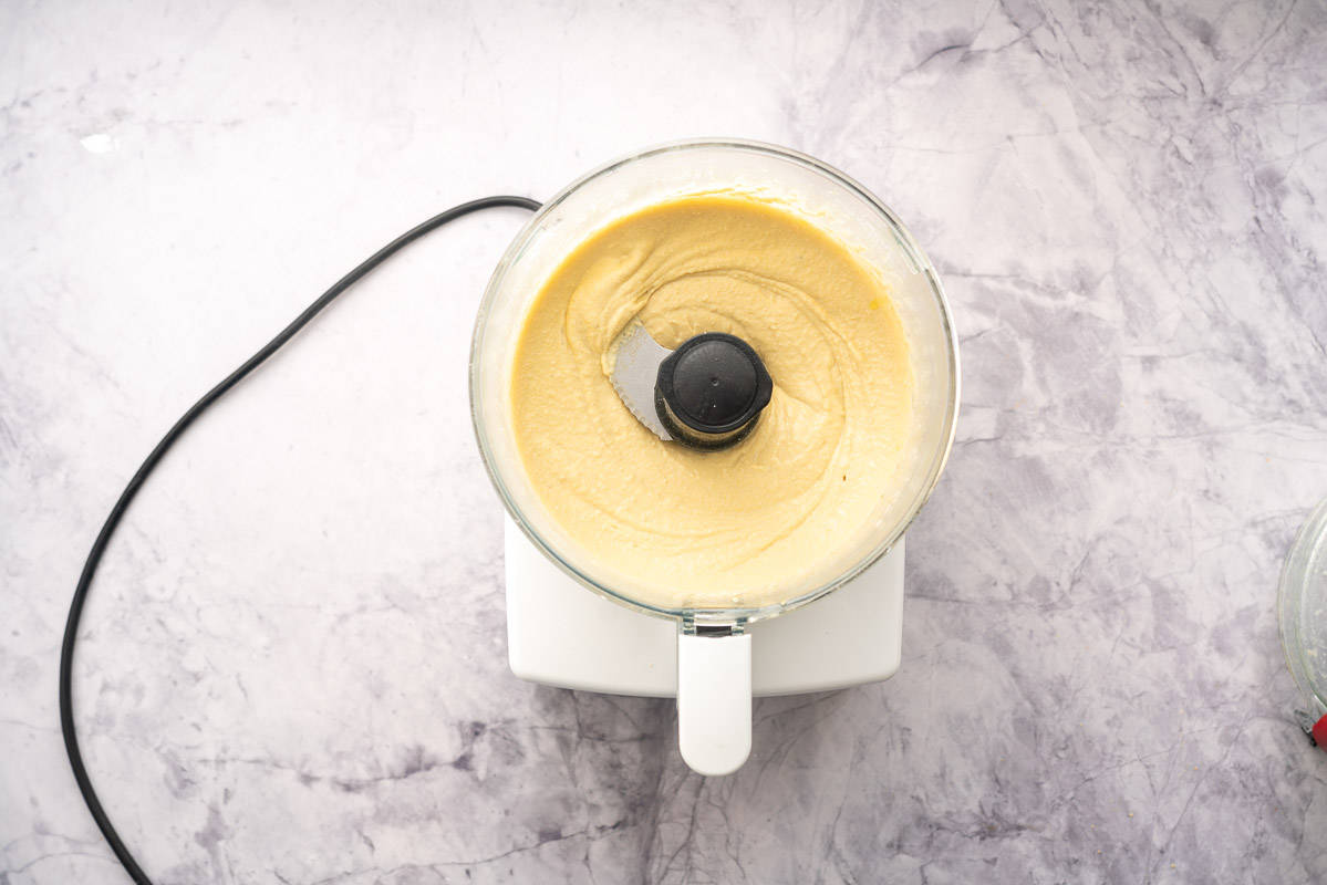 A food processor sitting on a bench with whipped hummus inside of it