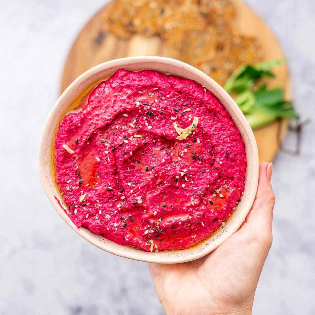 A hand holding up a bowl full of beetroot hummus, sprinkled with seasoning. 